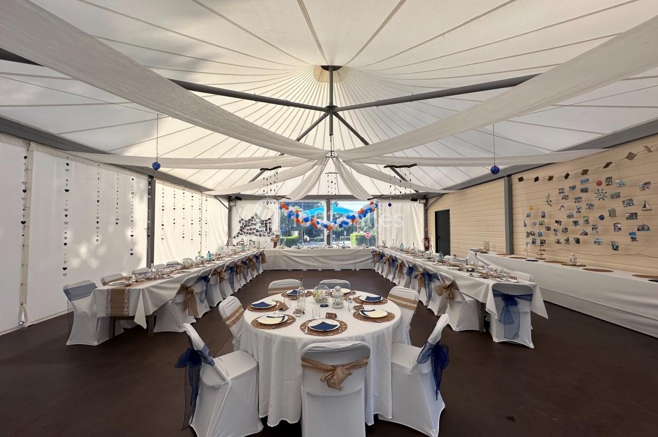 Salle de réception décorée avec des tables rondes dressées, chaises blanches ornées de nœuds bleus et plafond drapé.