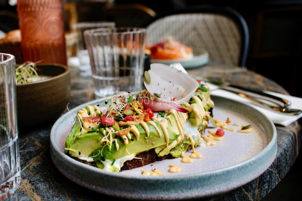 Toast garni d'avocat, graines, légumes frais et sauce, servi sur une assiette en céramique dans un restaurant.