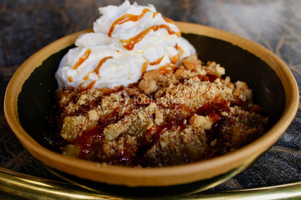 Dessert dans un bol en céramique avec crumble, crème fouettée et filet de caramel sur une table sombre.