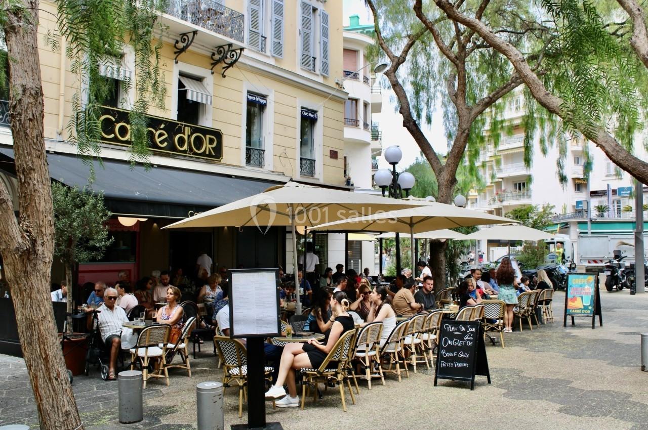 Terrasse animée d'un café en plein air, avec des clients assis sous des parasols dans une rue bordée d'arbres.
