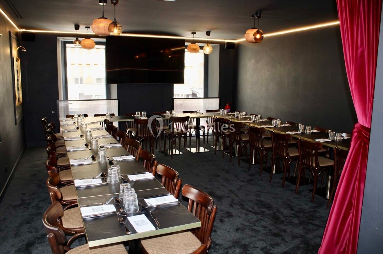 Salle de restaurant sombre avec deux longues tables dressées, chaises en bois et rideaux rouges encadrant l'entrée.