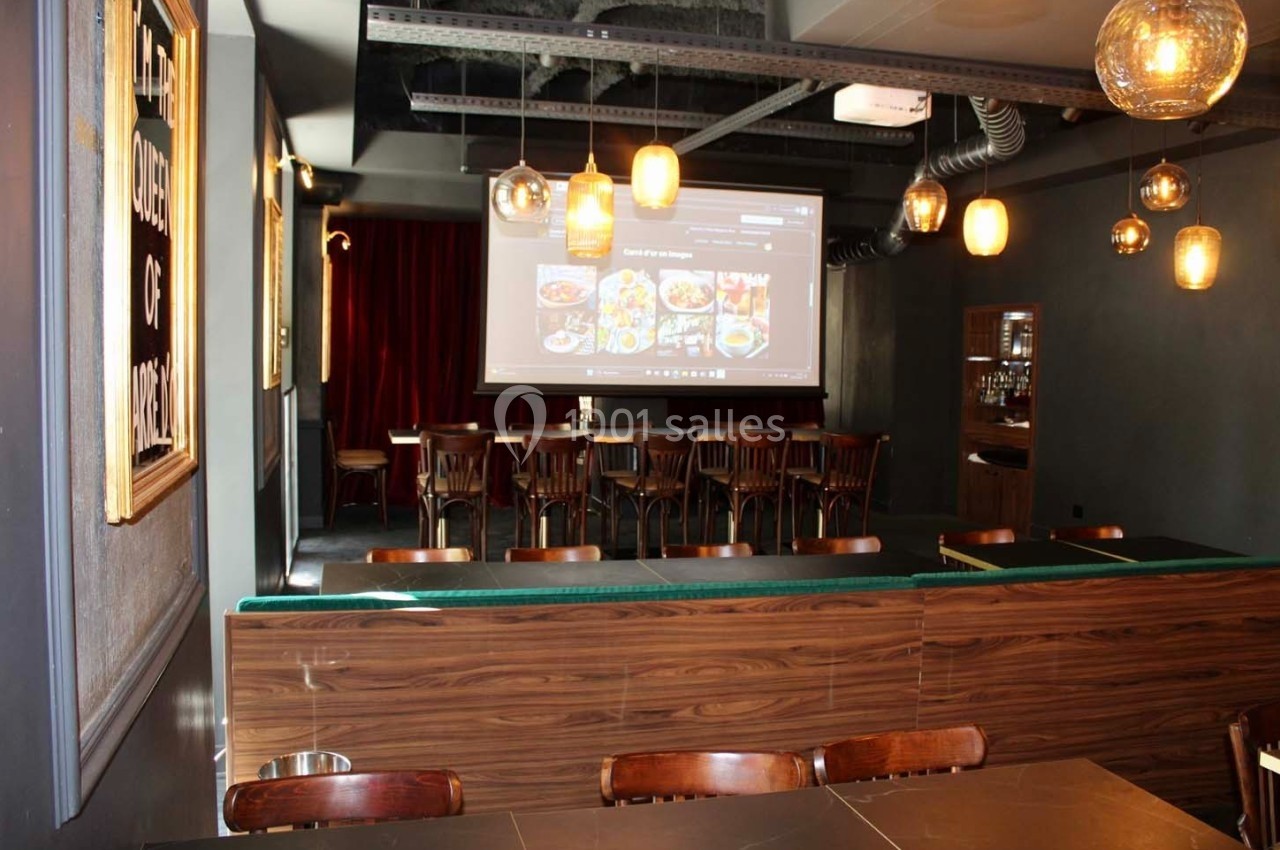Salle de restaurant avec tables en bois, chaises alignées, projecteur allumé et éclairage suspendu chaleureux.