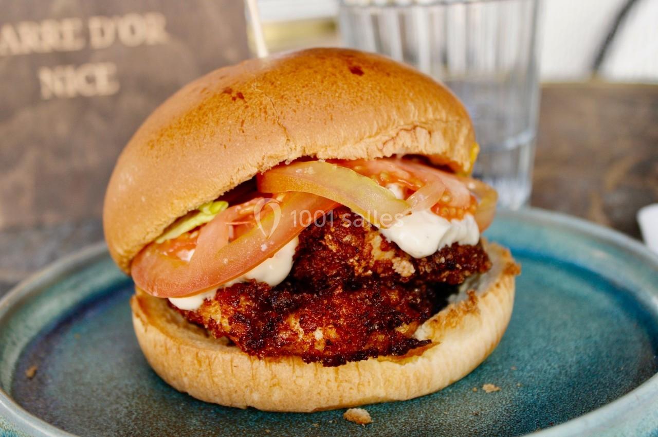 Burger avec poulet pané, tranches de tomate, salade et sauce, servi dans une assiette en céramique bleue.