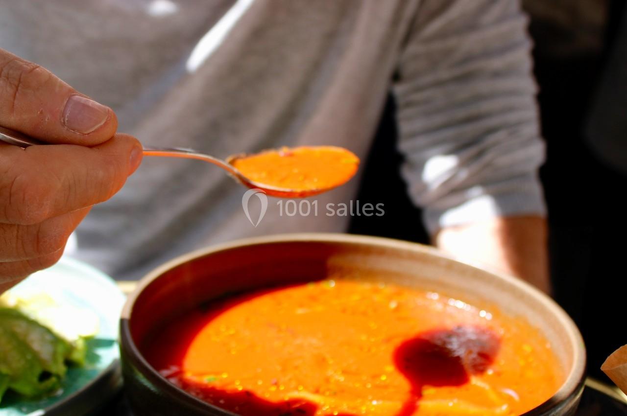 Une personne tient une cuillère remplie de soupe rouge au-dessus d'un bol en céramique, éclairée par la lumière naturelle.