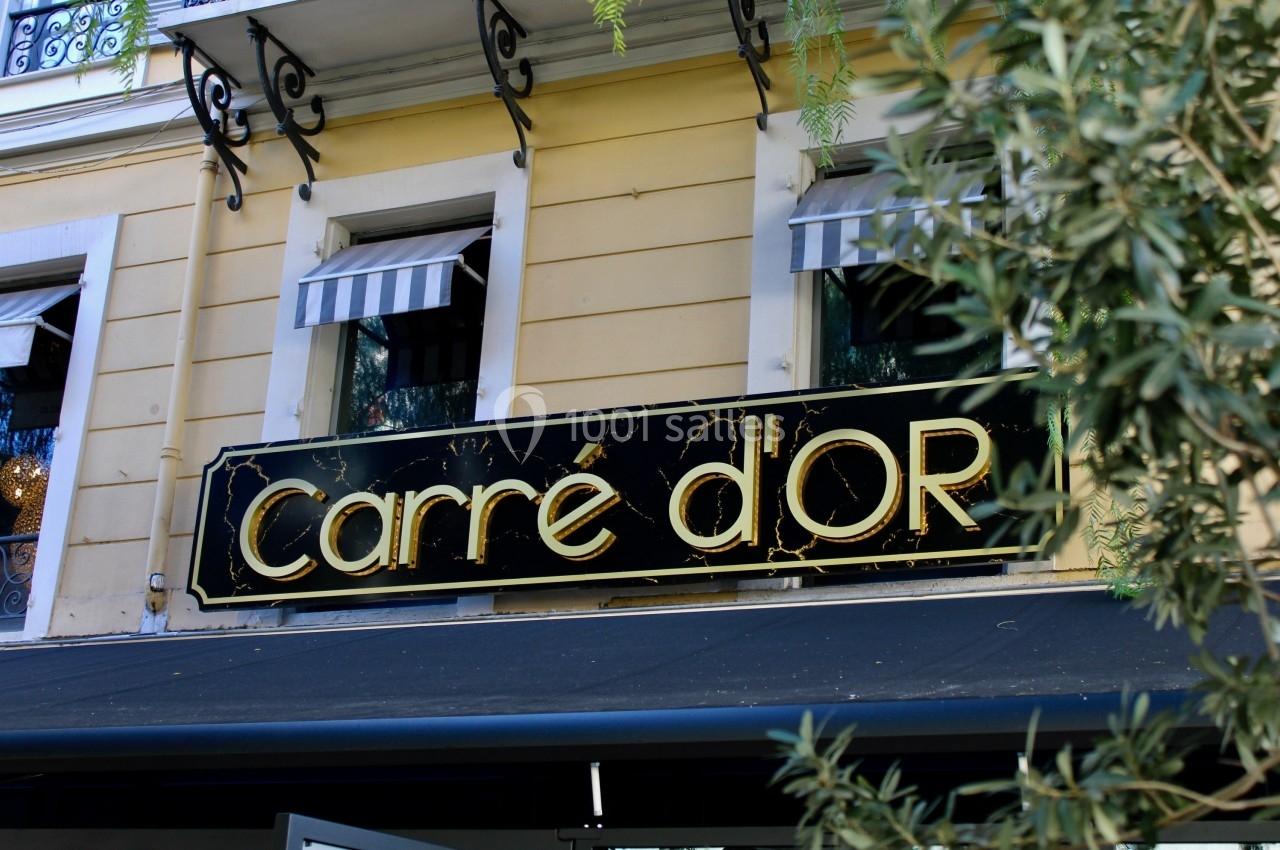Enseigne noire et dorée avec l'inscription ’Carré d'Or’ sur la façade d'un bâtiment jaune avec des stores rayés.