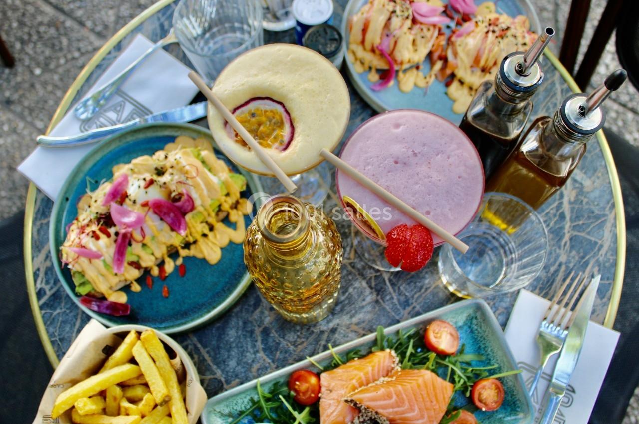 Table garnie de plats variés : saumon, frites, assiettes garnies, smoothies et condiments, sur une terrasse extérieure.