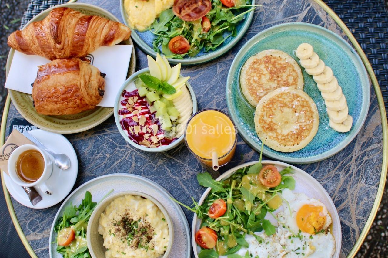 Petit-déjeuner varié avec croissants, œufs, pancakes, fruits, salade, café et jus d'orange sur une table ronde.