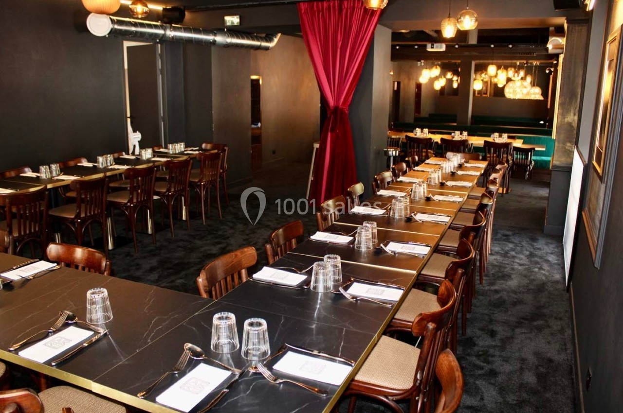 Salle de restaurant élégante avec longues tables noires, chaises en bois, rideaux rouges et éclairage chaleureux.