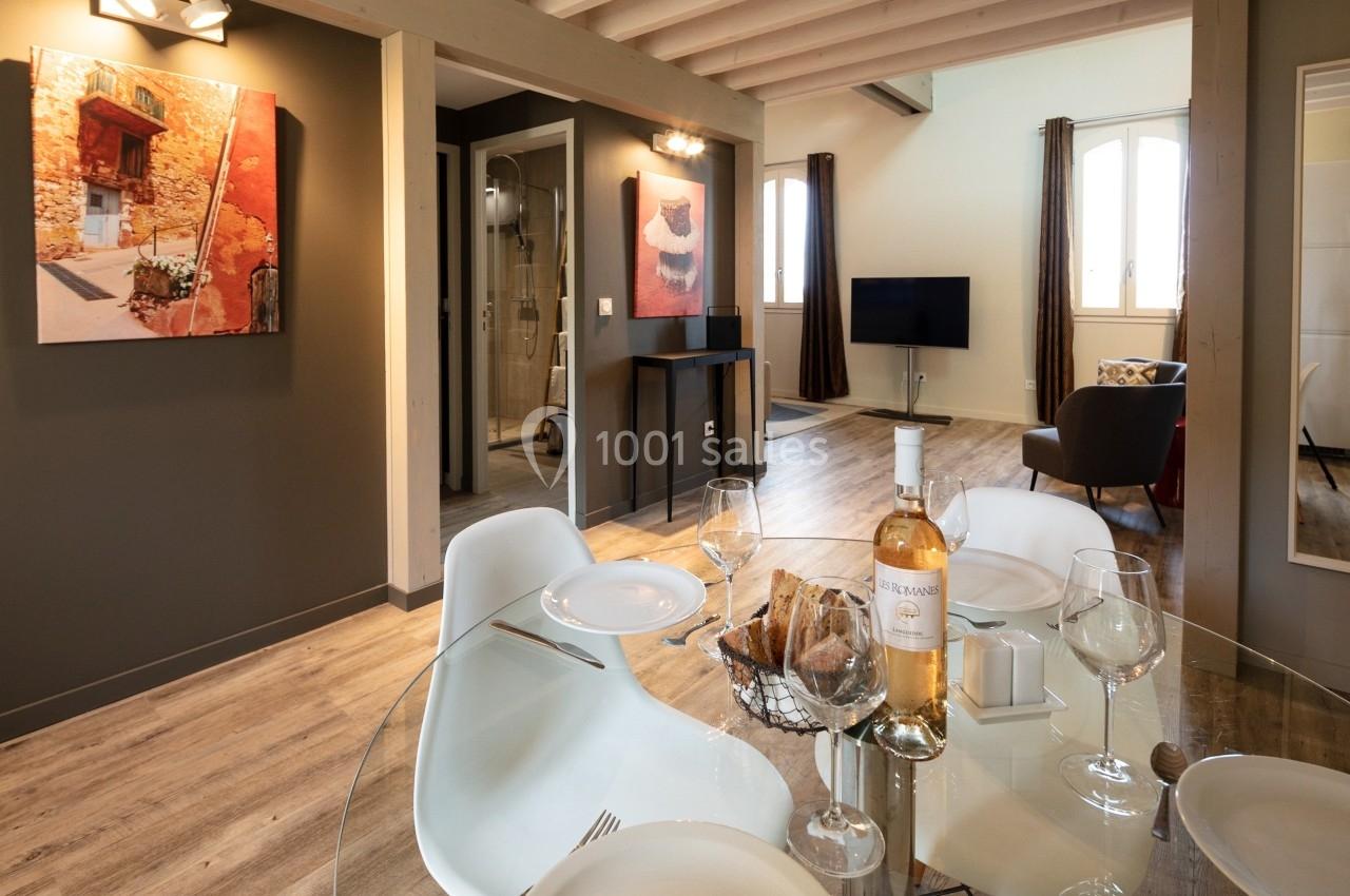 Salle à manger avec table en verre dressée, vue sur un salon moderne avec télévision et œuvres d'art au mur.