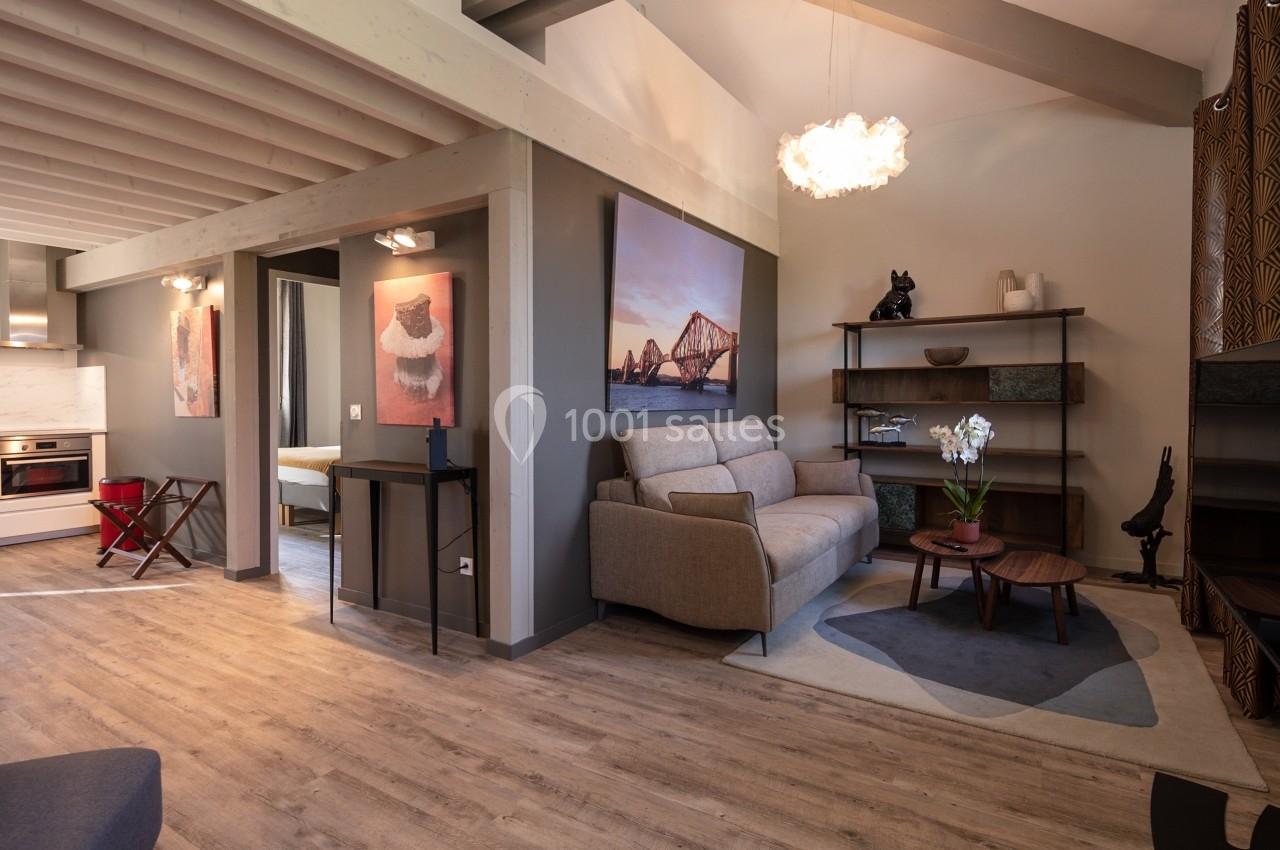 Salon moderne avec canapé gris, table basse en bois, étagères décoratives et vue partielle sur une cuisine ouverte.