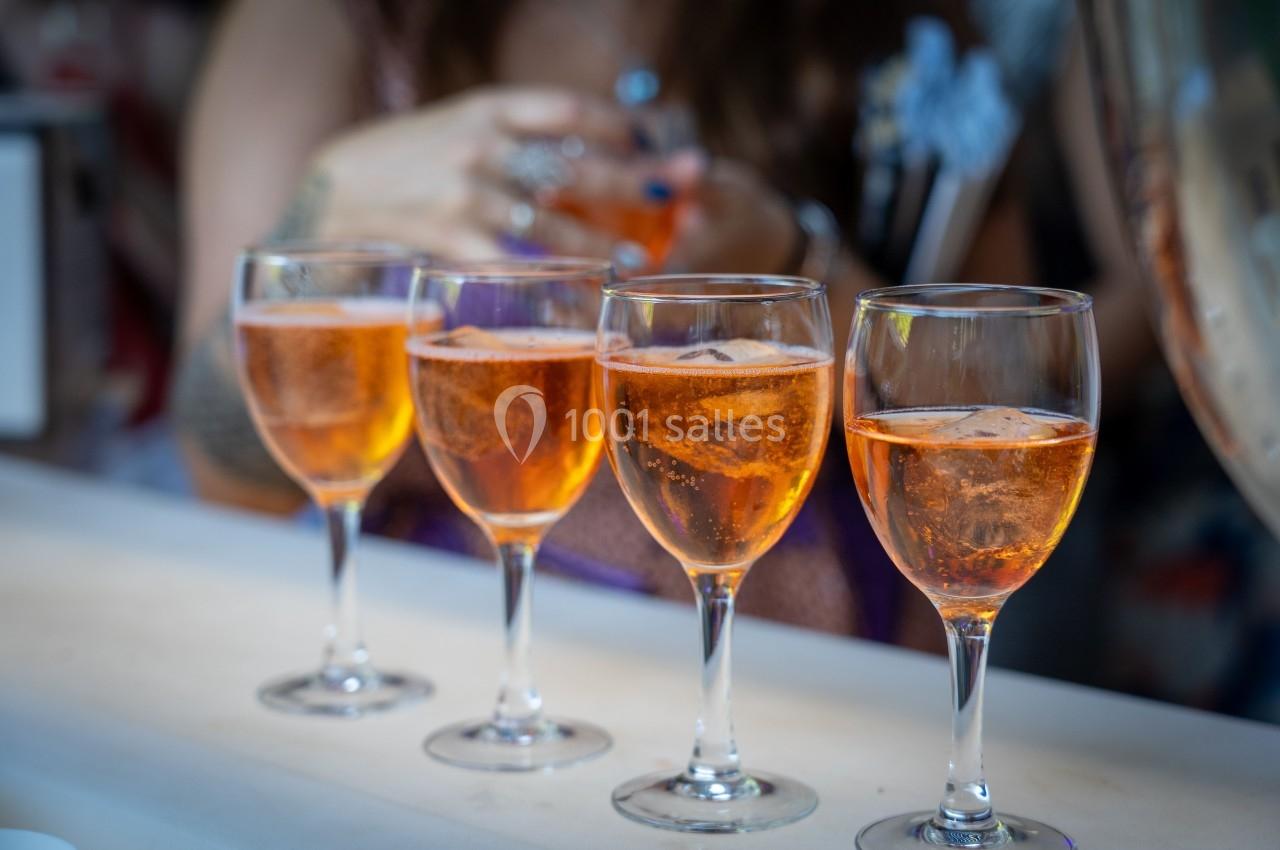 Quatre verres de cocktail orange alignés sur un comptoir, avec une personne floue en arrière-plan.