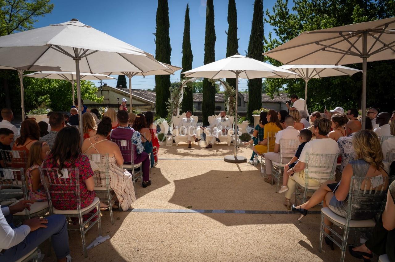 Cérémonie en plein air avec des invités assis sous des parasols, entourés d'arbres et d'un ciel bleu.