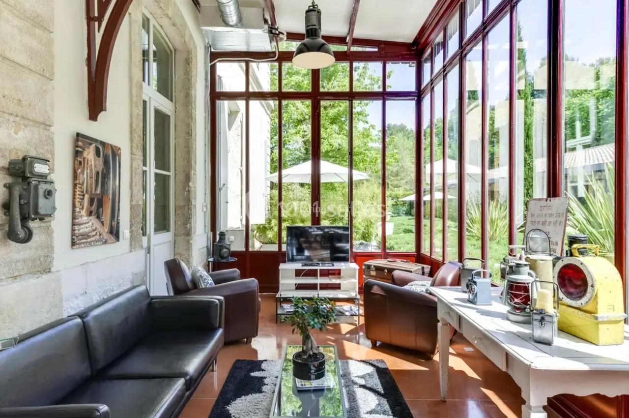 Véranda lumineuse avec murs vitrés, mobilier moderne, vue sur un jardin verdoyant et terrasse extérieure.