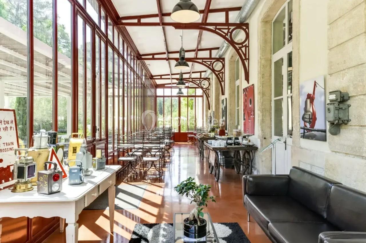 Salle lumineuse avec verrière, mobilier en métal et bois, canapés, et objets décoratifs sur des tables.