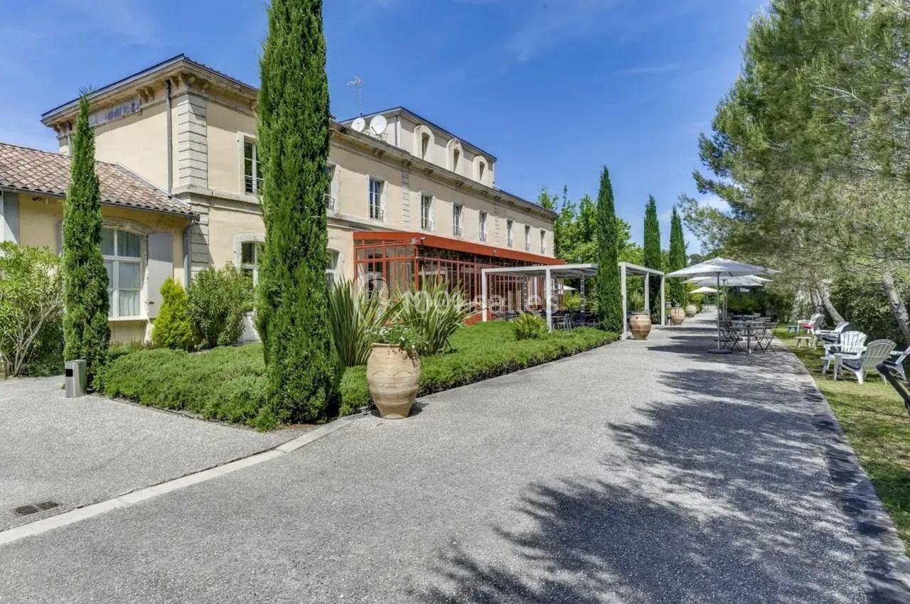 Allée bordée de végétation menant à une grande bâtisse en pierre avec terrasse et parasols, sous un ciel bleu.