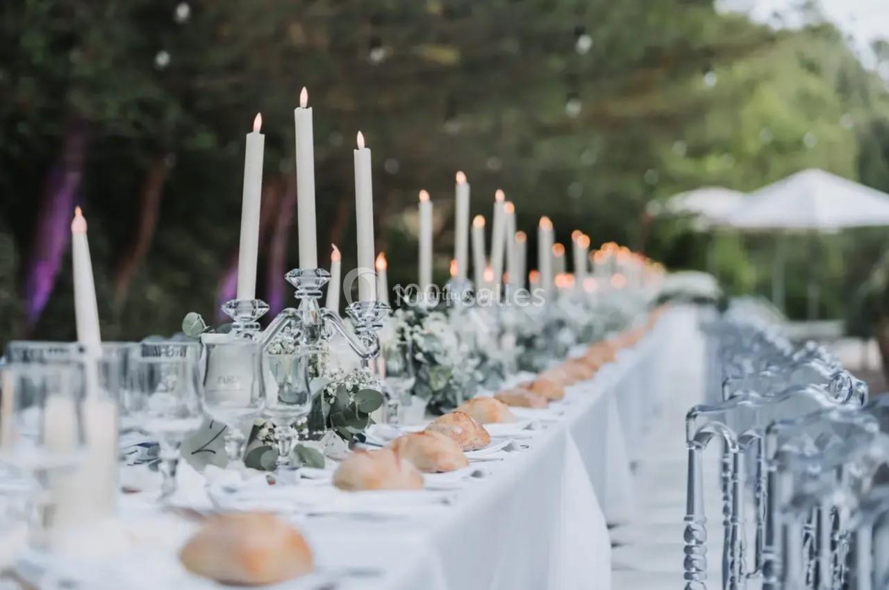 Grande table de réception extérieure décorée de chandeliers, fleurs blanches et vaisselle élégante.