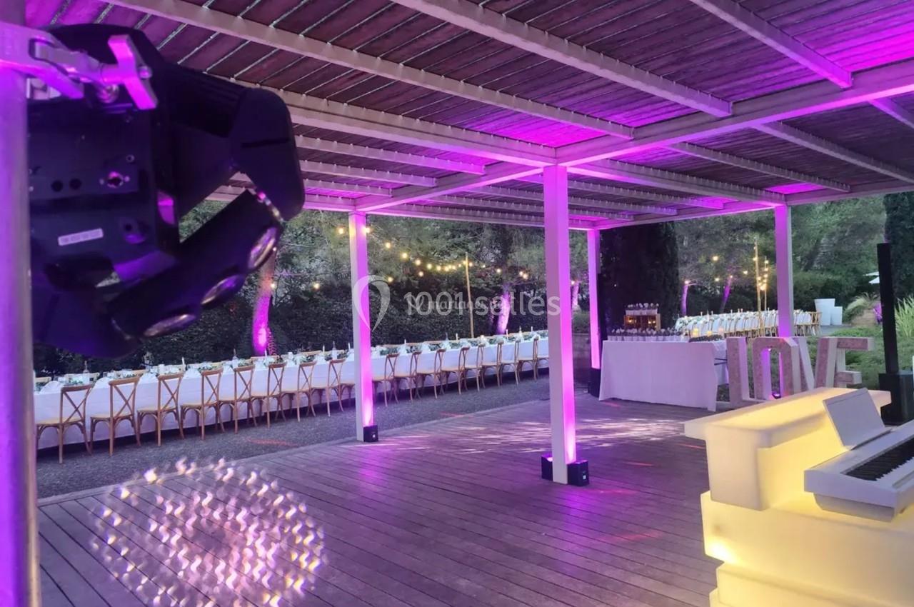 Terrasse en bois décorée de lumières violettes, avec tables dressées et projecteurs pour un événement en plein air.