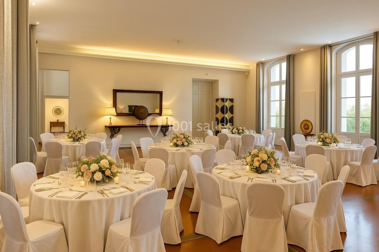 Salle de réception lumineuse avec des tables rondes dressées, nappes blanches et bouquets de fleurs au centre.