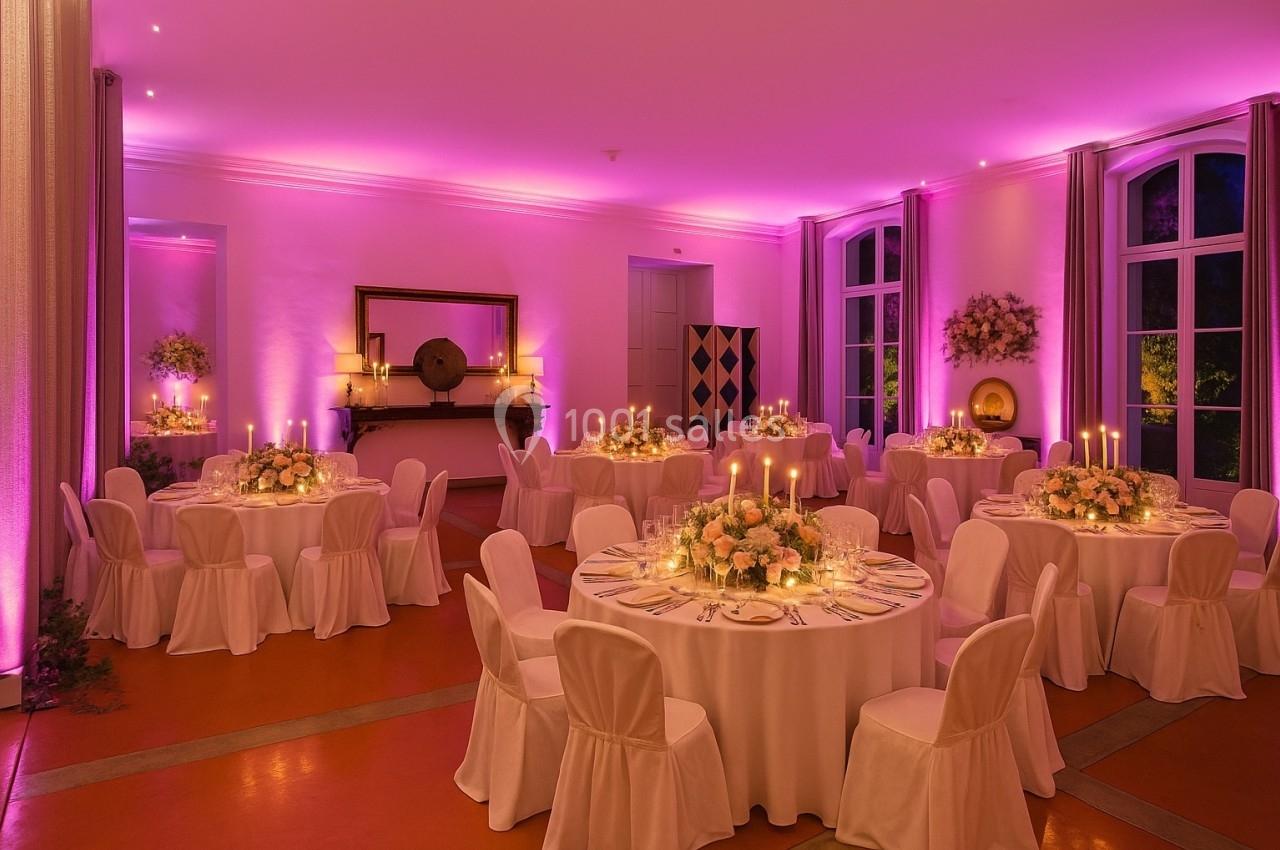 Salle de réception élégamment décorée avec des tables rondes, nappes blanches, bougies et éclairage rose tamisé.