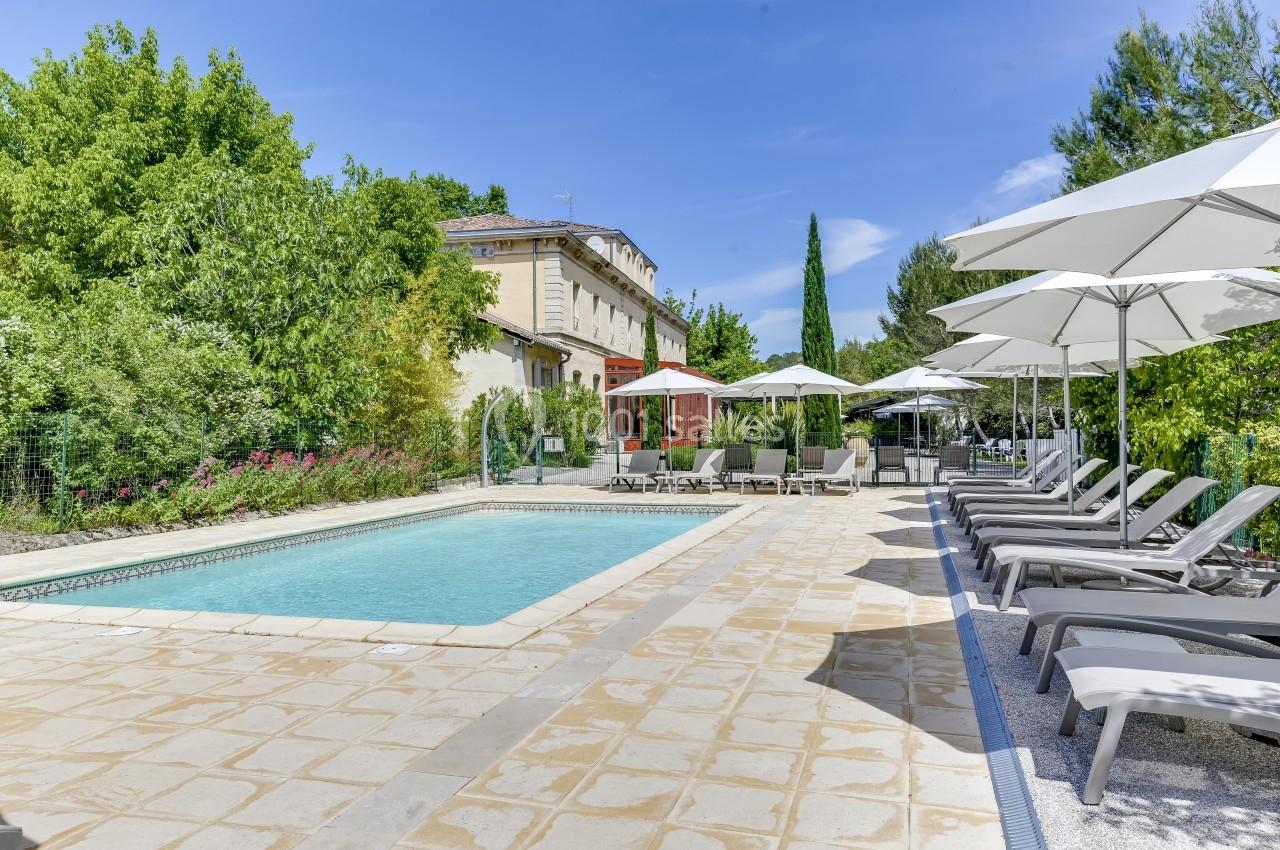 Piscine extérieure entourée de transats, parasols blancs et végétation, avec un bâtiment en arrière-plan.