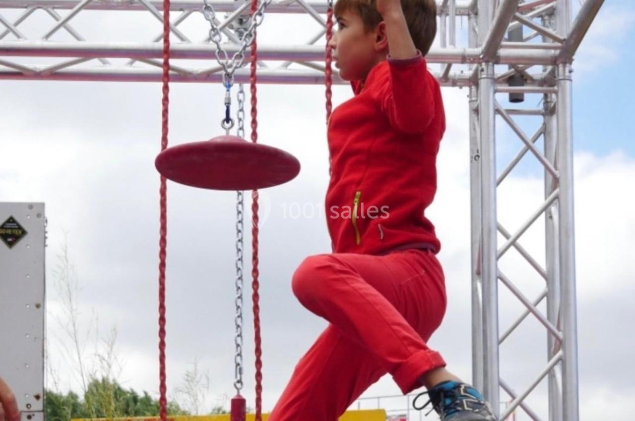 Un enfant en tenue rouge traverse un parcours suspendu en s'accrochant à une corde sous un ciel nuageux.