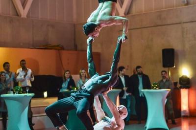 Deux acrobates exécutent une figure d'équilibre sur une caisse, devant un public dans un espace éclairé.