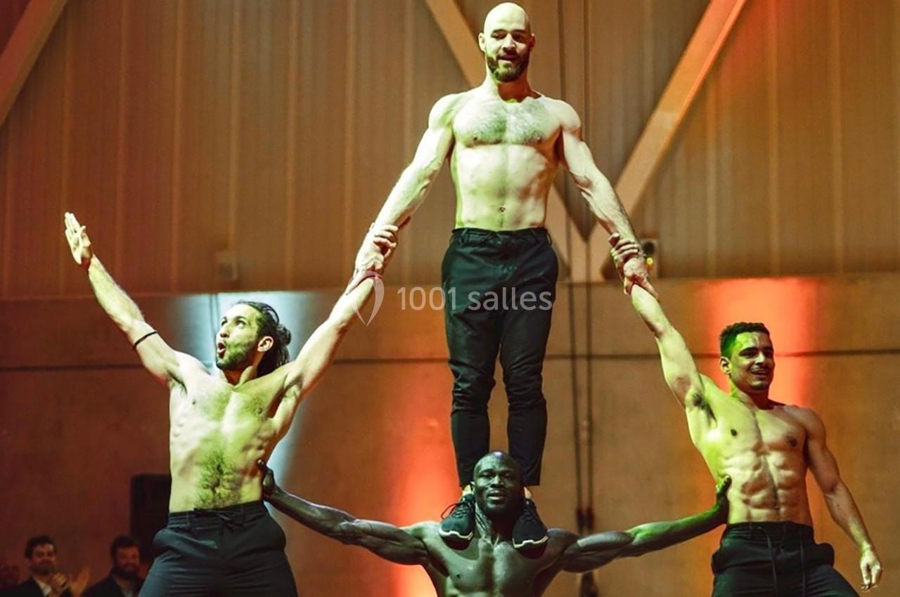 Quatre hommes réalisent une figure acrobatique en intérieur, avec un homme debout au sommet soutenu par les autres.