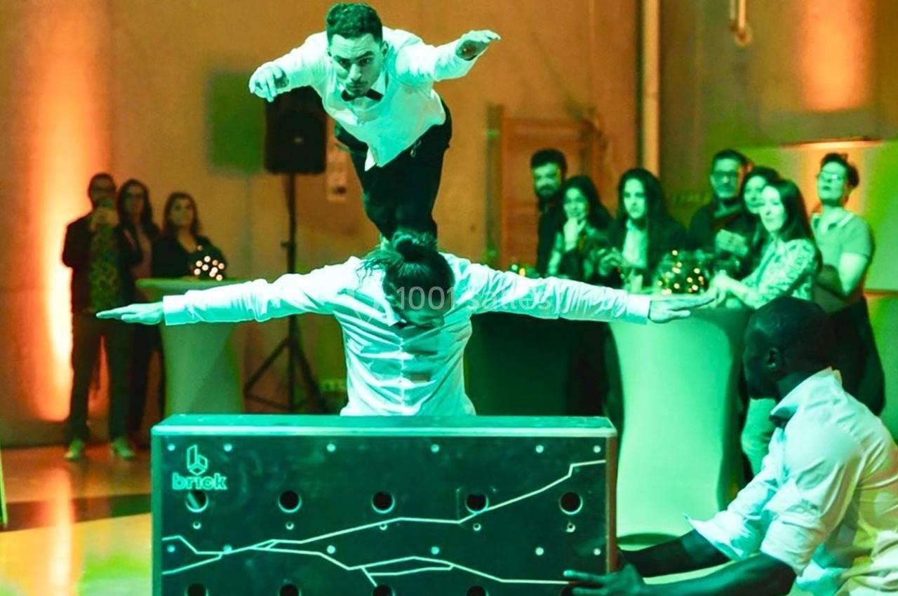 Deux acrobates exécutent une figure d'équilibre sur une caisse, devant un public dans un espace éclairé.