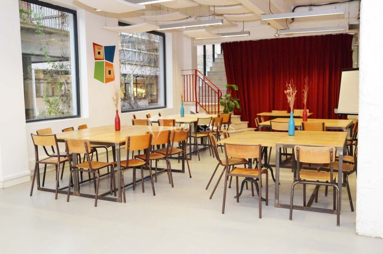 Salle lumineuse avec des tables en bois, des chaises, un rideau rouge en fond et des décorations murales colorées.