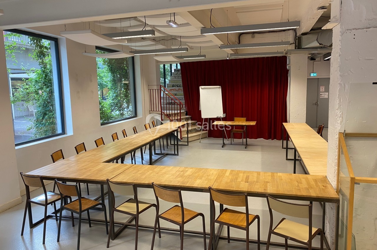 Salle de réunion avec tables disposées en U, chaises en bois, tableau blanc et rideau rouge en arrière-plan.