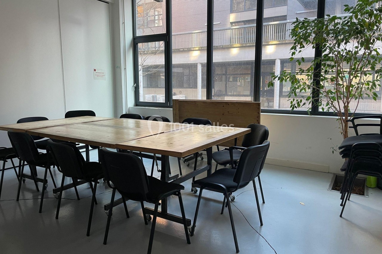 Salle de réunion lumineuse avec une grande table en bois entourée de chaises noires, près de fenêtres et d'une plante.