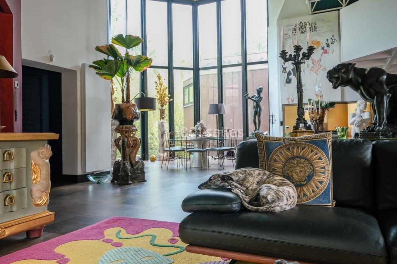 Salon lumineux avec grandes baies vitrées, mobilier éclectique, sculptures et chien allongé sur un canapé noir.