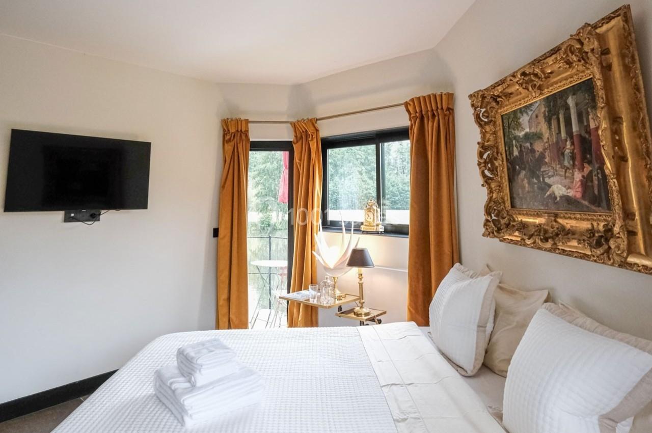 Chambre lumineuse avec lit double, draps blancs, télévision murale, fenêtres avec rideaux jaunes et tableau encadré.