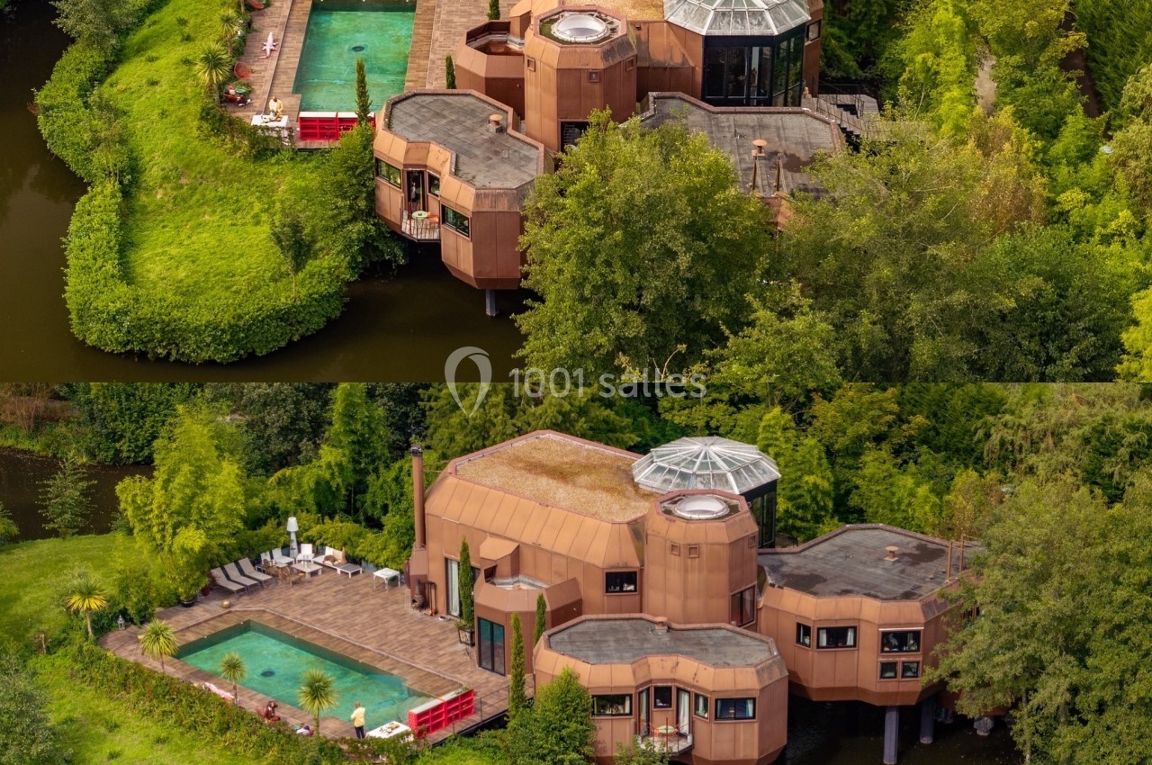 Vue aérienne d'une maison moderne entourée d'eau, avec piscine, terrasse et végétation luxuriante.