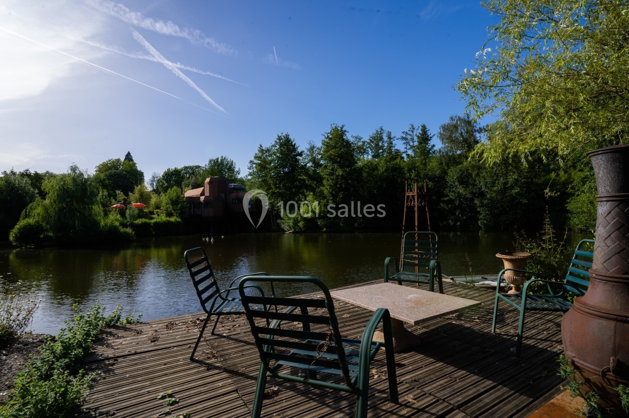 Terrasse en bois avec des chaises autour d'une table, donnant sur un étang entouré de végétation et d'arbres.
