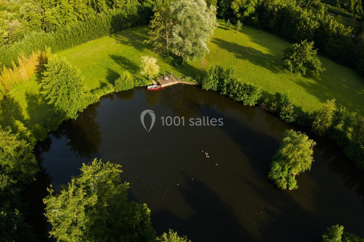 Vue aérienne d'un étang entouré de verdure avec un petit ponton, une barque rouge et des arbres en arrière-plan.