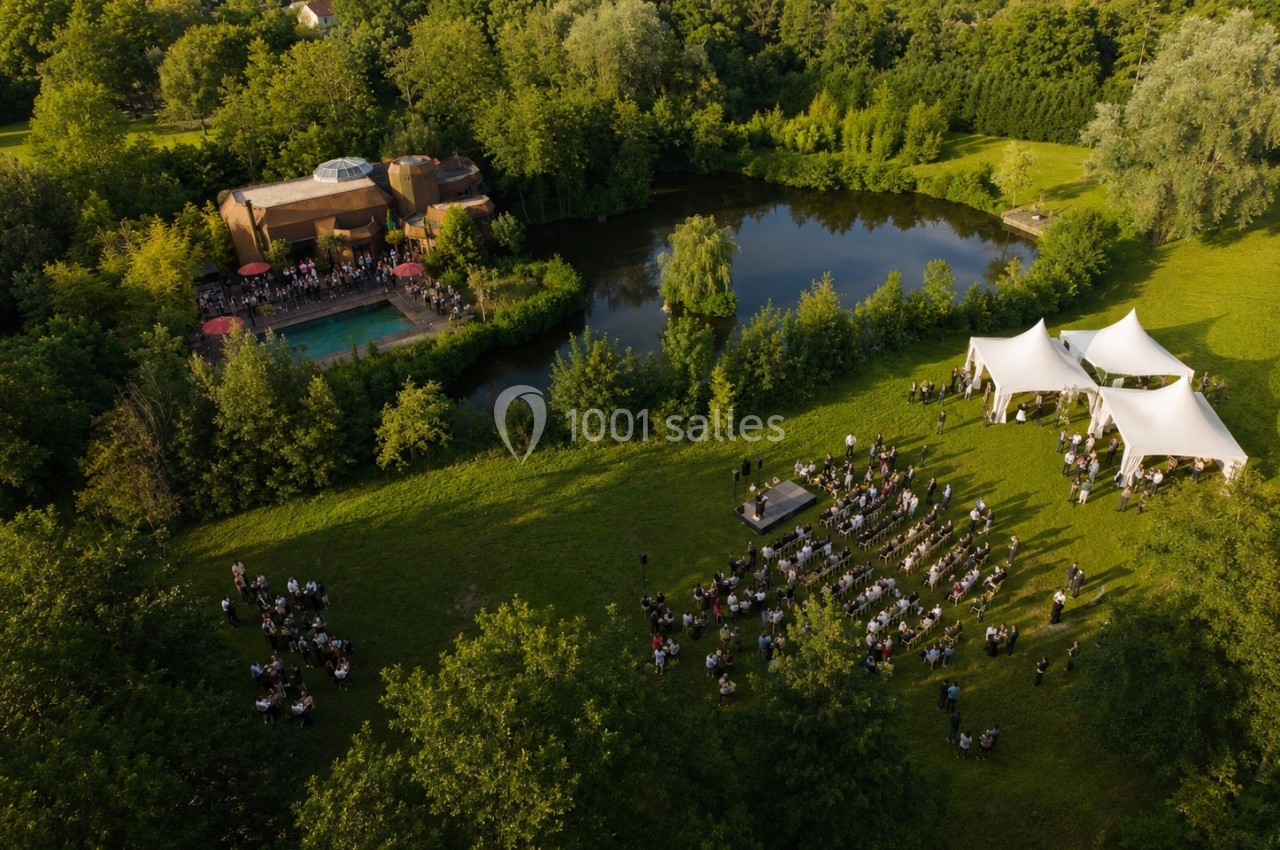 Vue aérienne d'un événement en plein air avec invités, tentes blanches, scène et étang entouré de verdure.