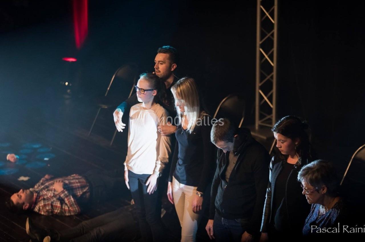 Un groupe de personnes debout sur une scène sombre, éclairées par un projecteur, avec une personne allongée au sol.