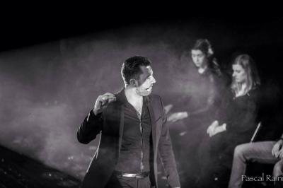 Un homme barbu pose devant un fond vert avec des cartes de trèfle flottant et des faisceaux lumineux.