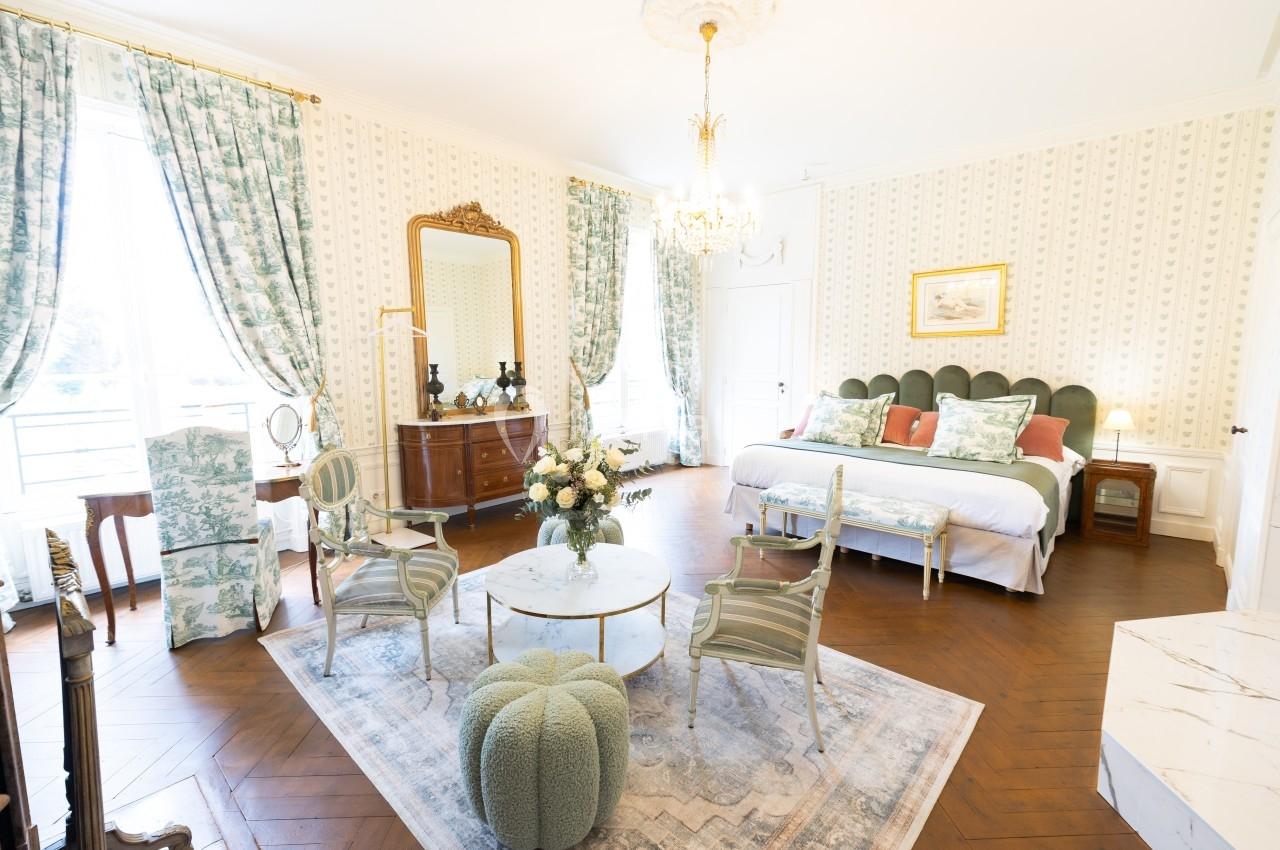 Chambre lumineuse avec lit double, mobilier classique, tapis, rideaux fleuris et lustre, dans un style élégant.
