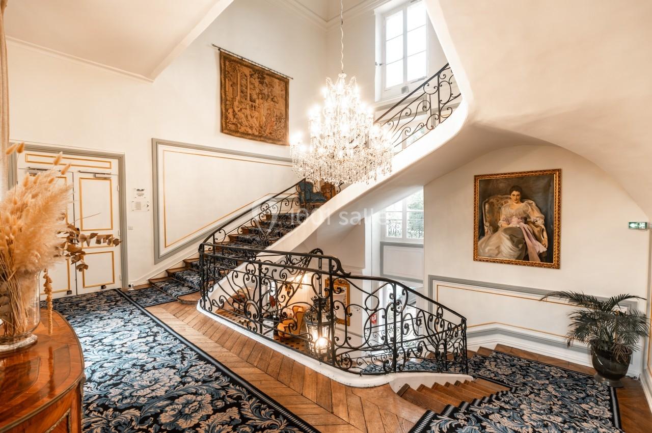 Escalier intérieur élégant avec tapis bleu, rampe en fer forgé, lustre imposant et tableaux décoratifs sur les murs.