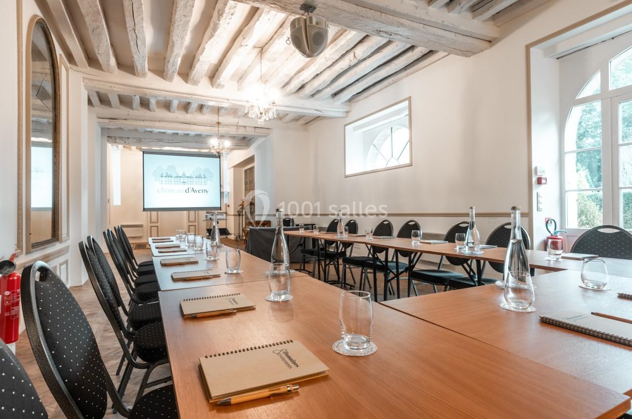 Salle de réunion lumineuse avec tables en U, chaises noires, bouteilles d'eau et écran de présentation.