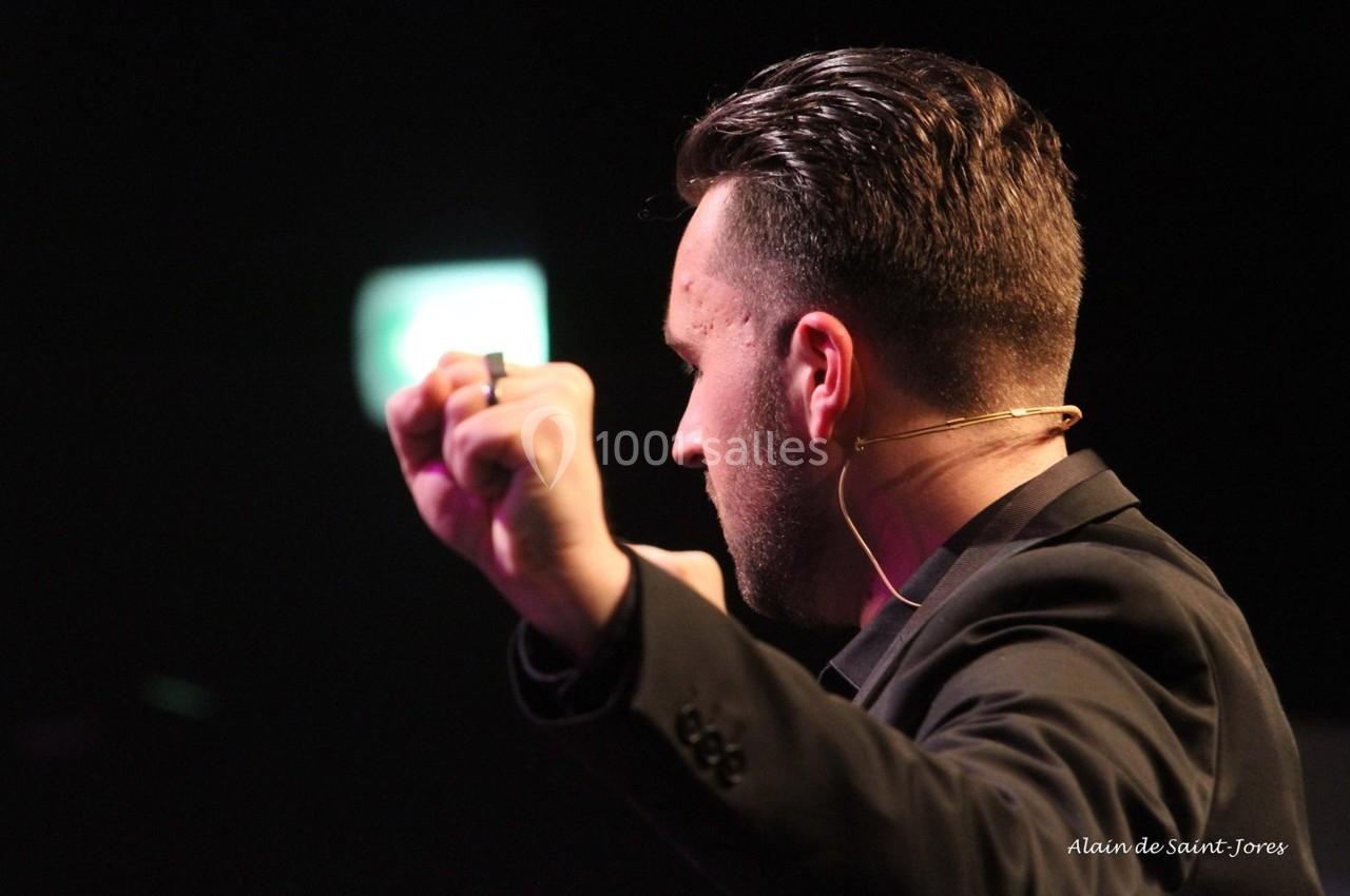 Homme de dos en costume noir, parlant avec un micro casque, les bras levés, dans un environnement sombre.