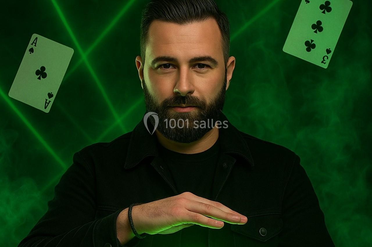 Un homme barbu en noir pose devant un fond vert avec des cartes à jouer et des faisceaux lumineux.