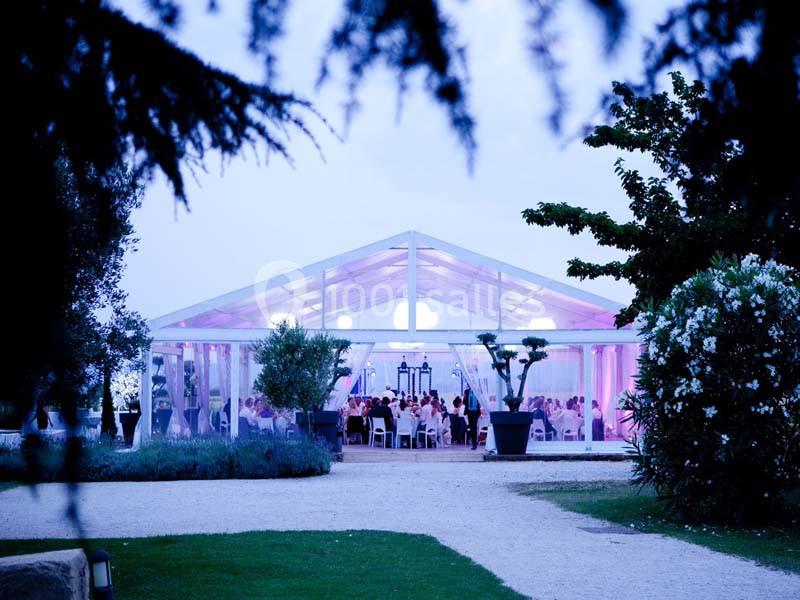 Vue d'une réception en soirée sous une tente transparente éclairée de lumières roses, entourée de verdure.