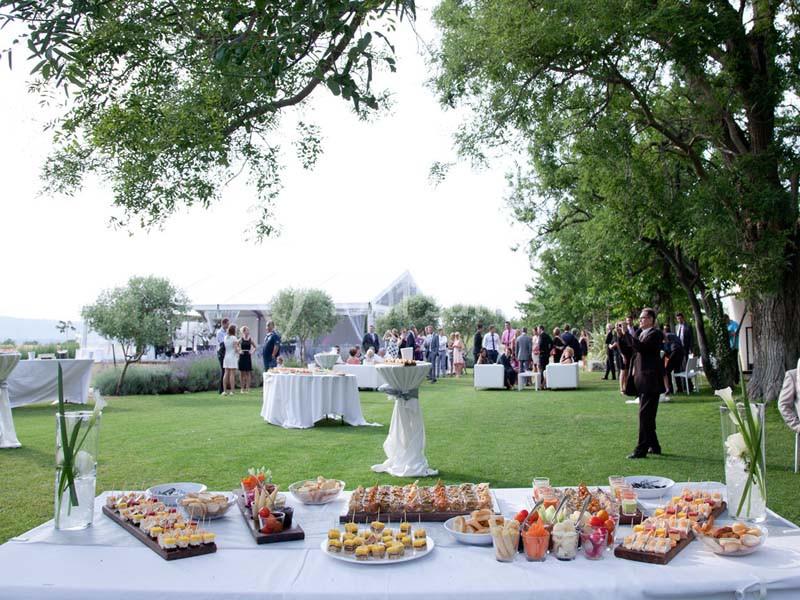 Table garnie de plats et desserts lors d'un événement en plein air, avec des invités rassemblés en arrière-plan.