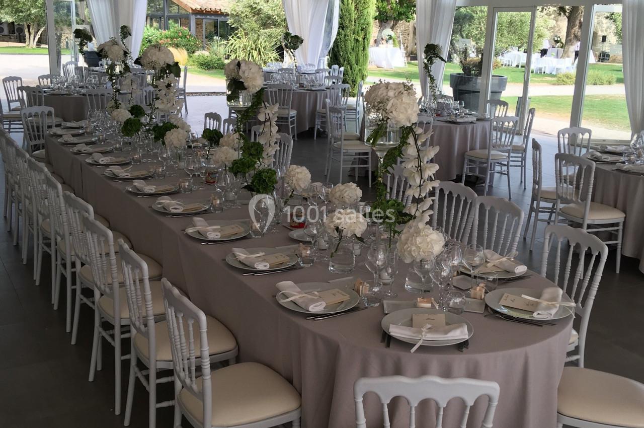 Salle de réception élégante avec tables dressées, nappes beiges, chaises blanches et décor floral sous un plafond drapé.