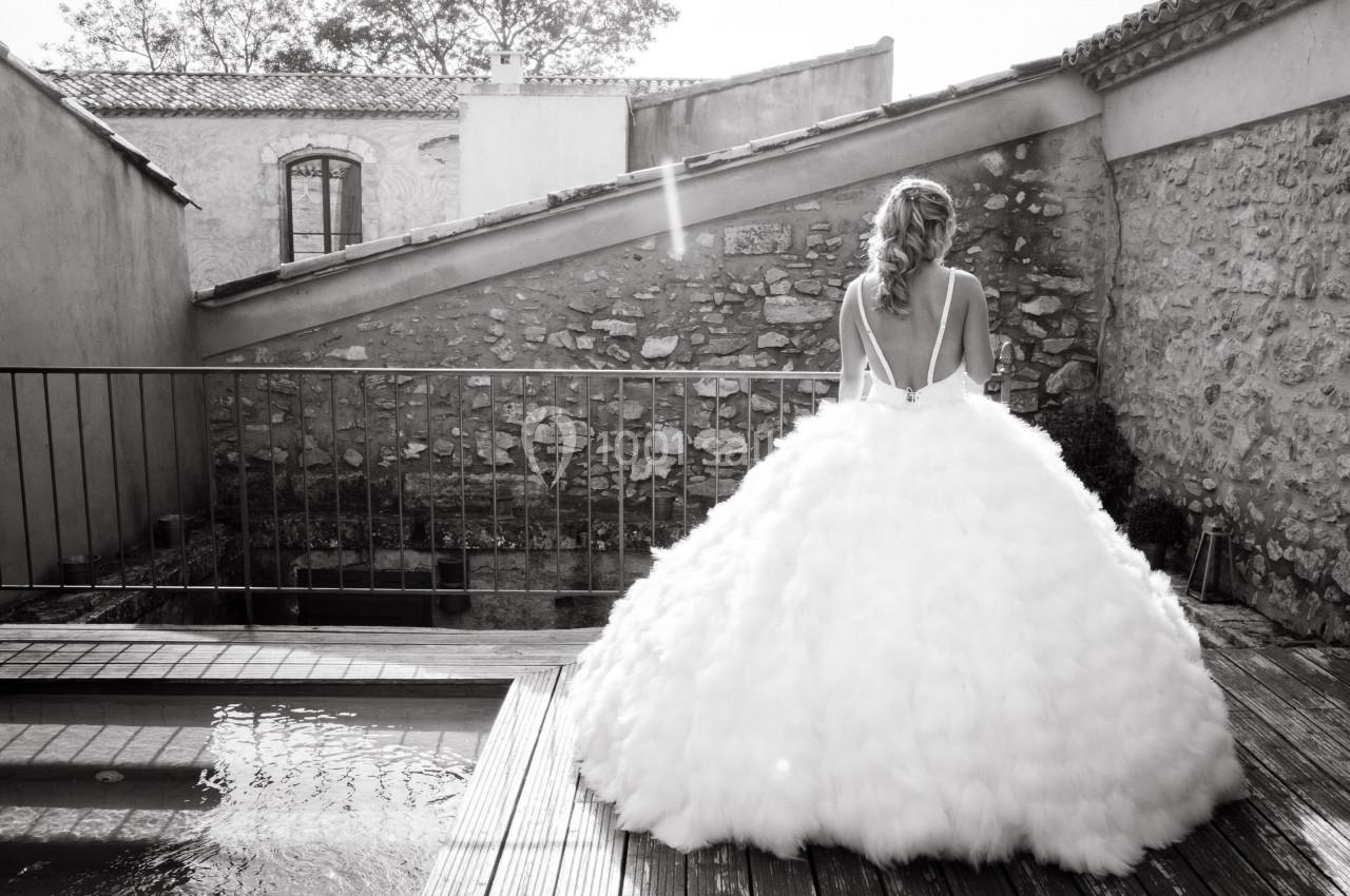 Une femme en robe de mariée volumineuse vue de dos, debout sur une terrasse en bois près d'une petite piscine.