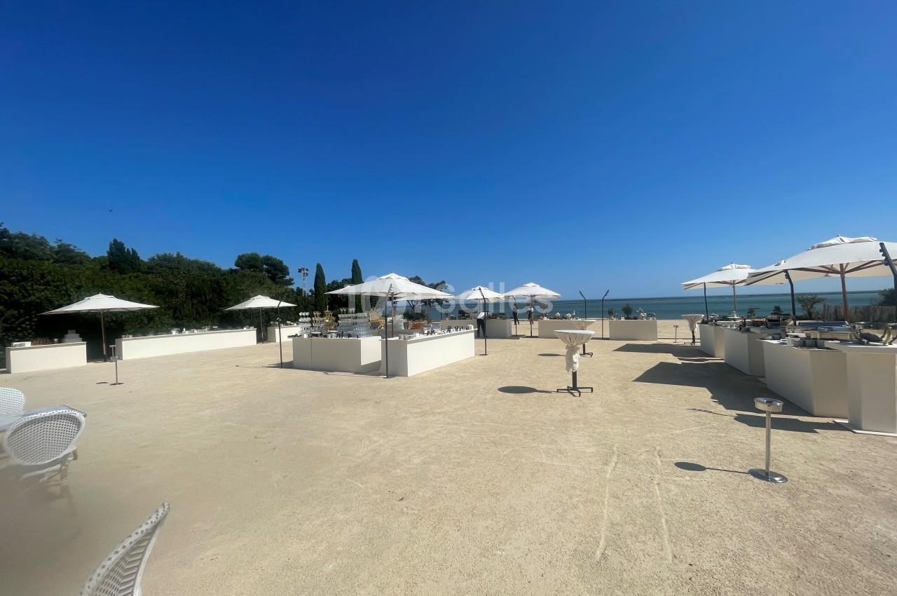Terrasse en plein air avec parasols blancs, mobilier clair et vue sur la mer sous un ciel bleu dégagé.