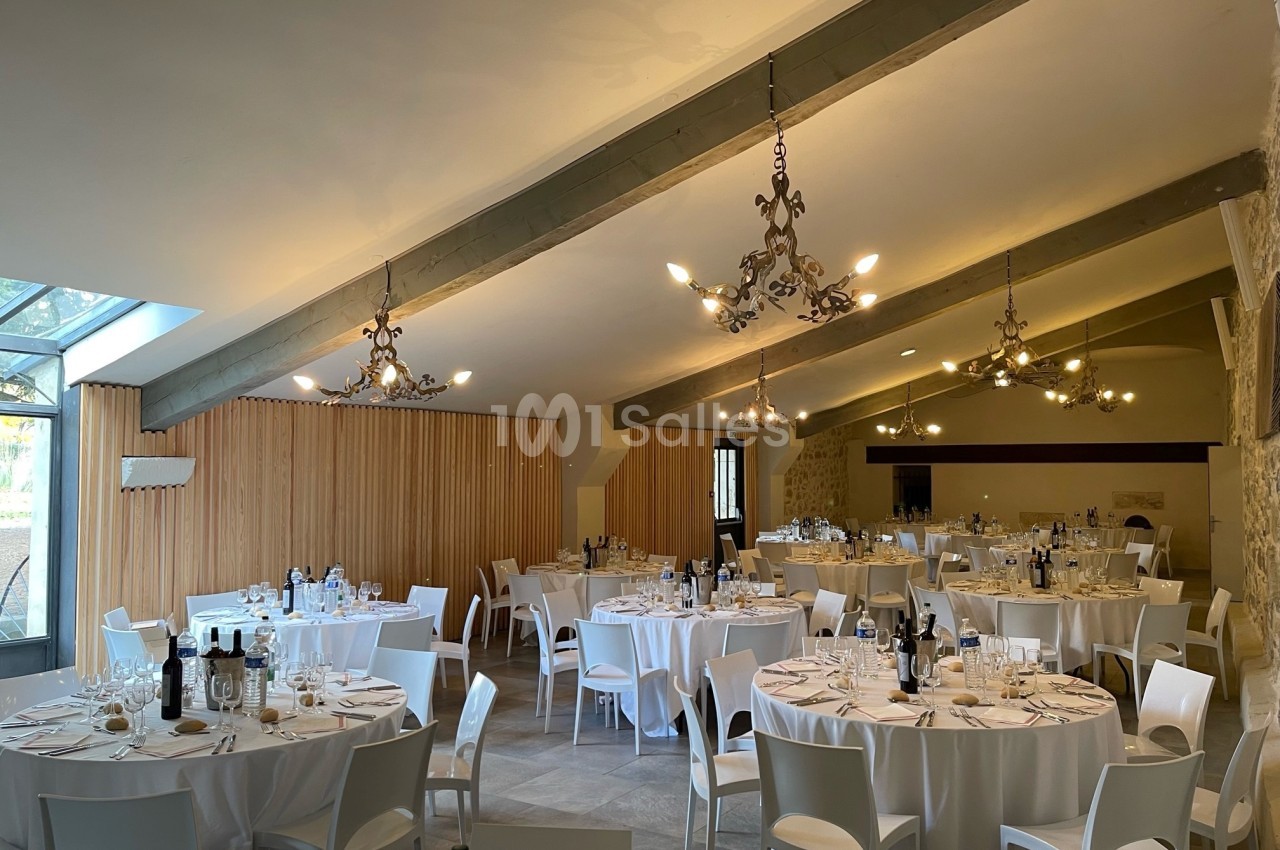 Salle de réception lumineuse avec tables rondes dressées, nappes blanches et chaises modernes sous un plafond avec poutres…